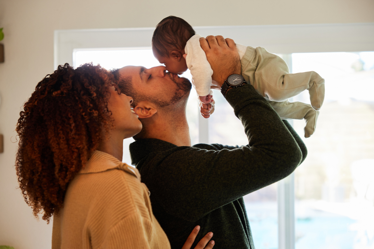 Mother with dark curly hair looking at her husband as he lifts and kisses their infant