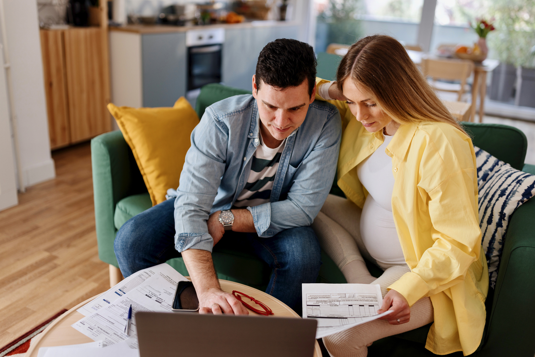 A pregnant woman and her partner reviewing documents and using a laptop, likely researching the cost to get a breast pump. The image reflects planning and budgeting for baby-related expenses.