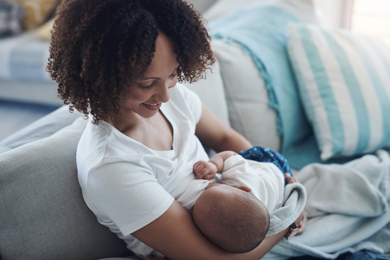 African American woman breastfeeding her infant on a couch after giving birth while contemplating how soon to pump or breastfeed