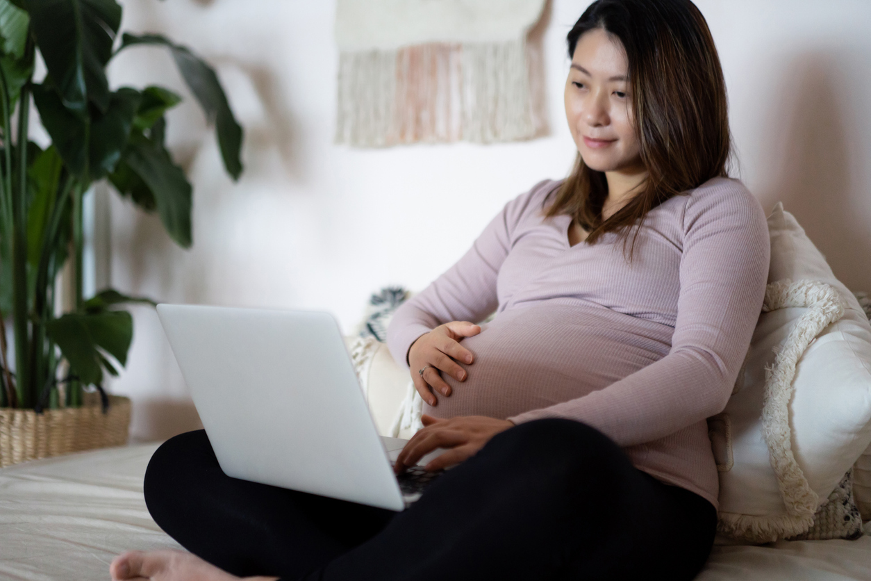 Pregnant woman in purple shirt sitting on bed looking at laptop ordering a breast pump through insurance