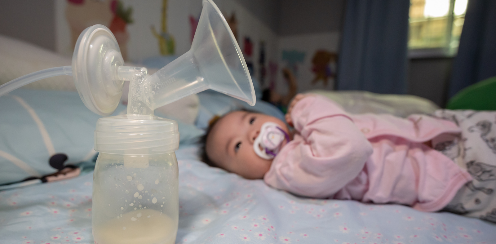 A baby lying on a bed with a pacifier, next to a bottle and breast pump, illustrating how new mothers can access a breast pump through Medicaid for support in their breastfeeding journey.