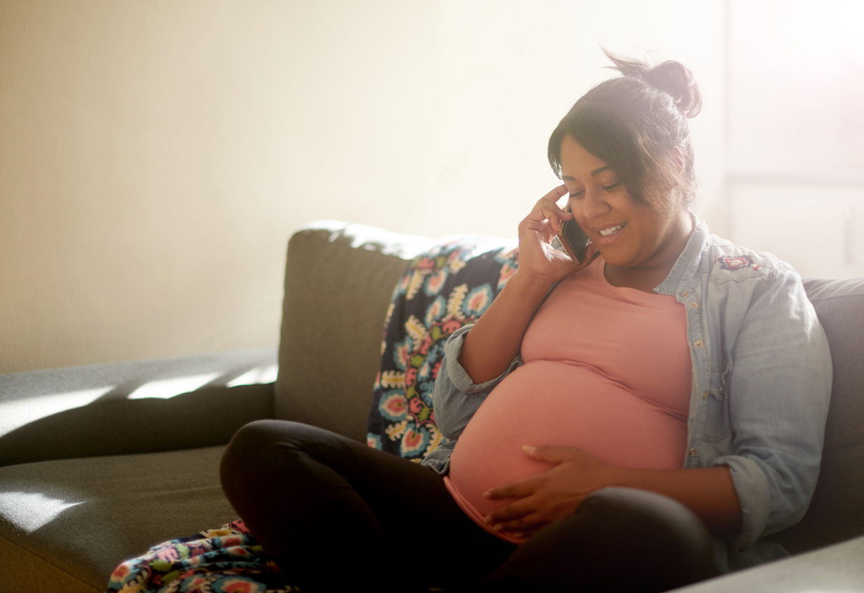 Pregnant woman sitting on the couch while on the phone obtaining a free breast pump through insurance