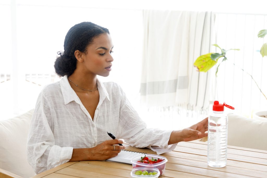 A woman sitting at a table with a notebook, planning and organizing with healthy snacks and water nearby. This image illustrates prep tips to keep in mind before breast pumping, emphasizing the importance of hydration, nutrition, and planning for a successful pumping session.