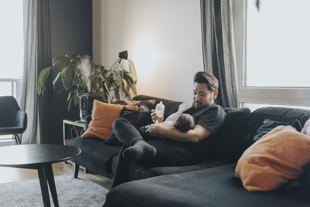 A father sitting on a couch, feeding a newborn with a bottle, showcasing the benefits of pumping. This image highlights how pumping allows other family members to bond with and nourish the baby.