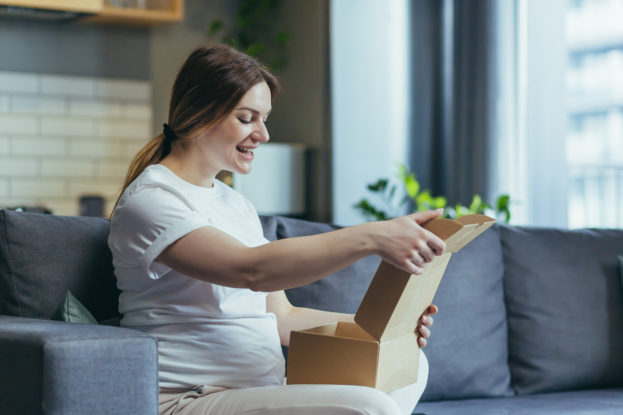 Pregnant woman in a white t-shirt sitting on a gray/blue couch opening her breast pump after ordering a breast pump through insurance