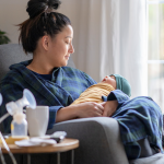 A new mother sitting on a rocking chair in her living room wearing a housecoat holding her newborn baby. There is a Spectra breast pump and nursing supplies around.