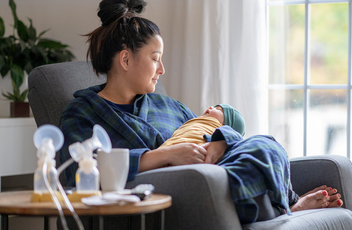 A new mother sitting on a rocking chair in her living room wearing a housecoat holding her newborn baby. There is a breast pump and nursing supplies around.