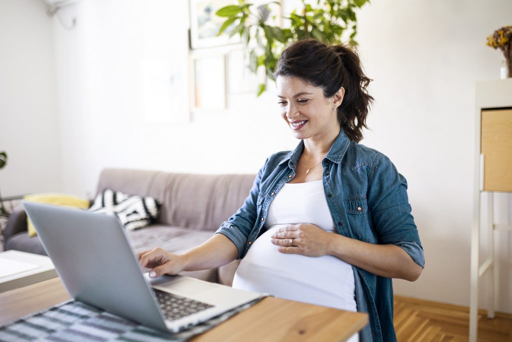 A pregnant woman smiling while researching on her laptop, possibly reading about breast pumps. This image represents breaking down Willow Pumps and exploring features to help expectant mothers make informed decisions.